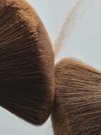 Two makeup brushes colliding mid-air, releasing a fine cloud of loose cosmetic powder against a pale background