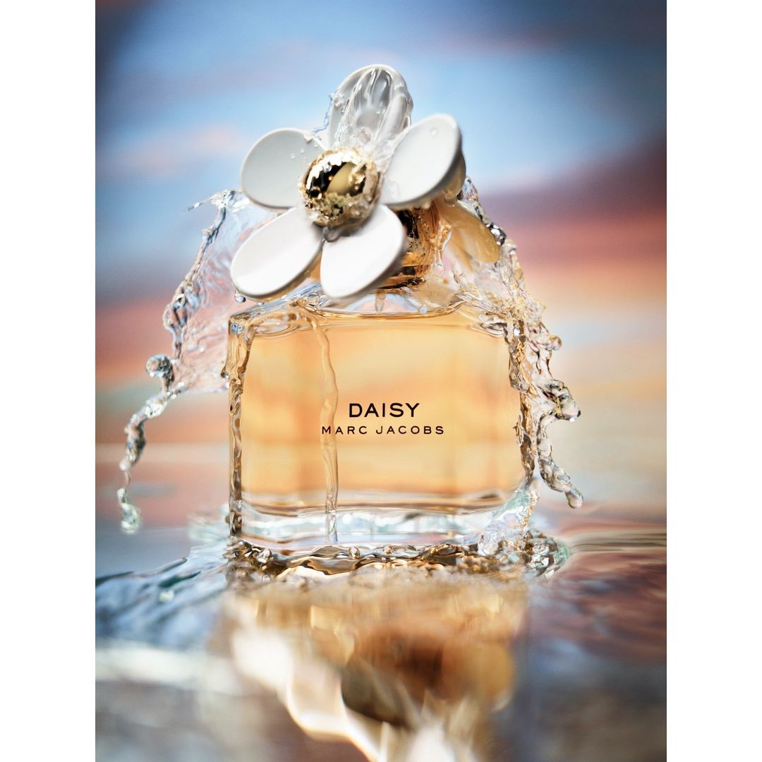 Product shot of Marc Jacobs Daisy perfume bottle with a white daisy cap and gold center, surrounded by splashing water against a blue background.