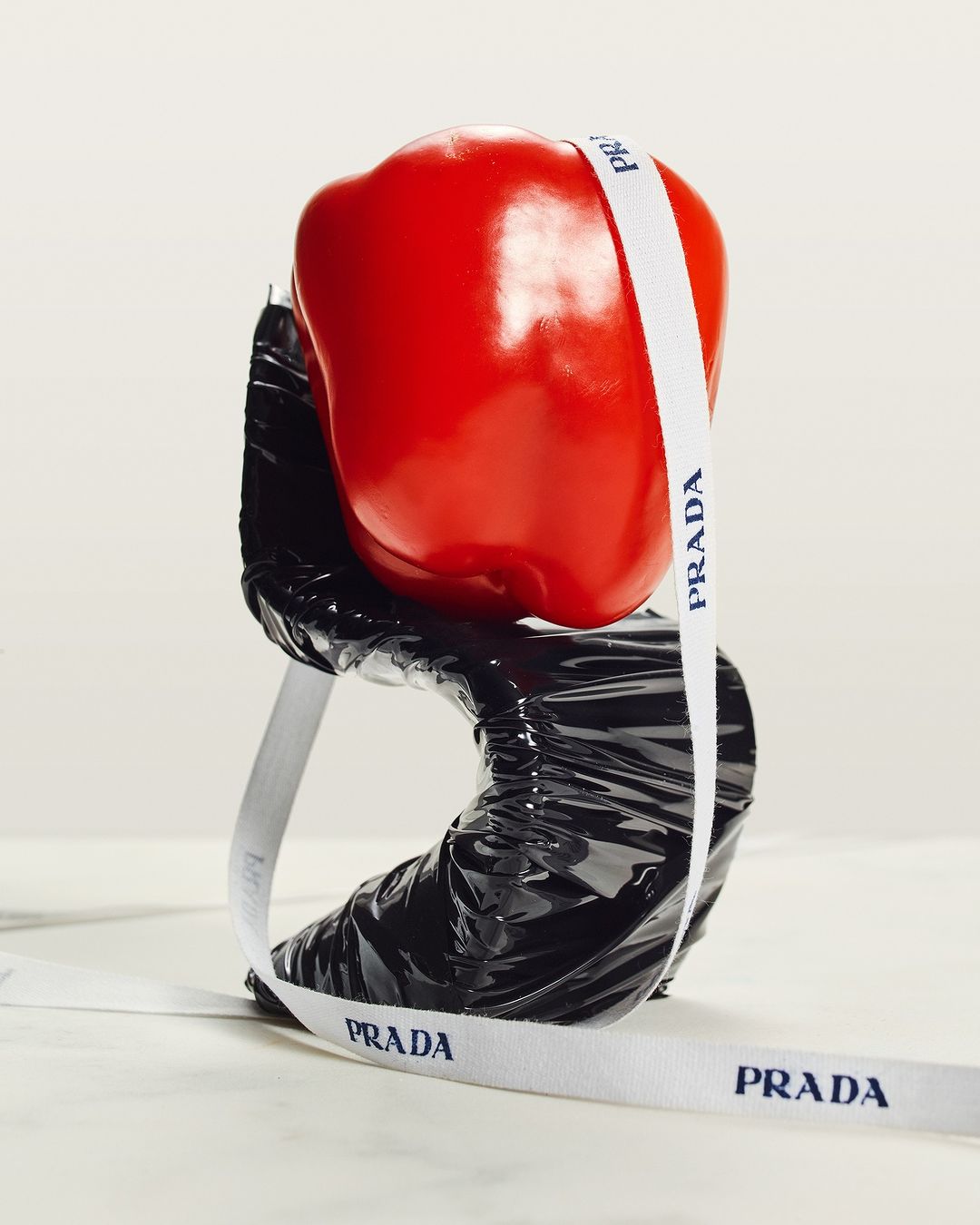 Still life food photography featuring a red bell pepper wrapped with a Prada branded ribbon, resting on a black glossy surface on a white background.