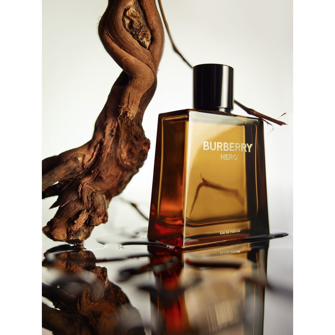 A glass Burberry Hero fragrance bottle with a dark brown cap sits next to a twisted wood branch with a reflective foreground surface.