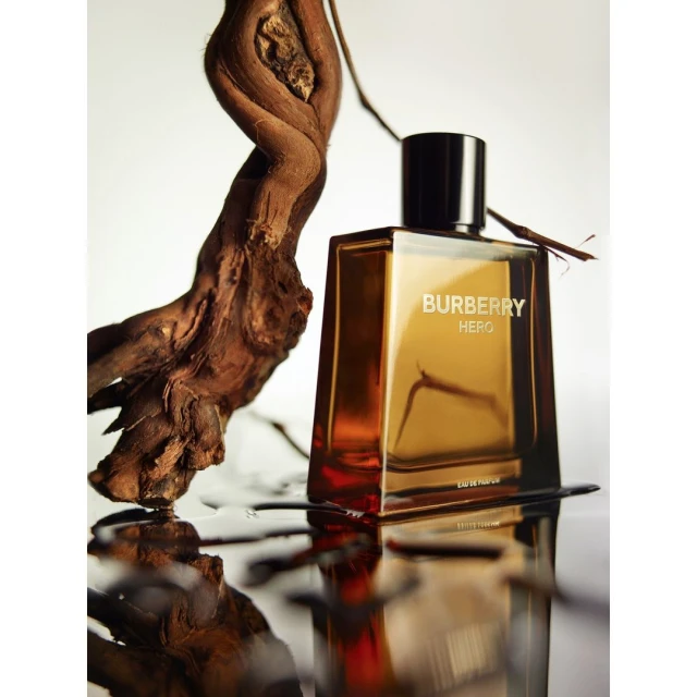 A glass Burberry Hero fragrance bottle with a dark brown cap sits next to a twisted wood branch with a reflective foreground surface.