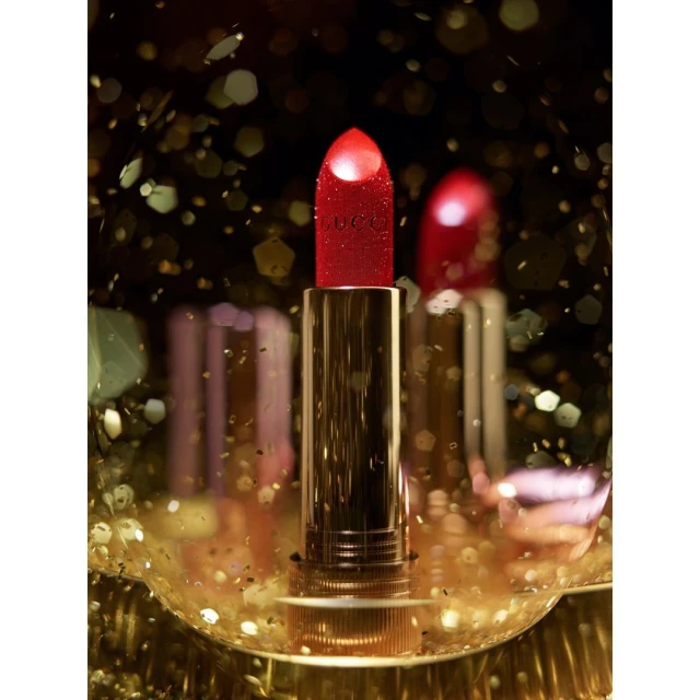 Close-up studio shot of two red and gold Gucci lipsticks surrounded by blurry golden bokeh for a beauty advertisement.