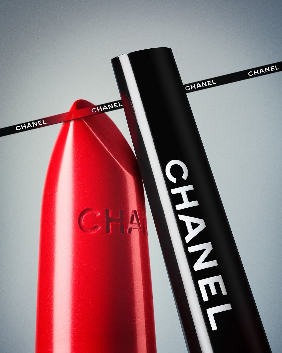 Close-up of a red Chanel lipstick alongside its black cap against a grey gradient background with branded ribbon