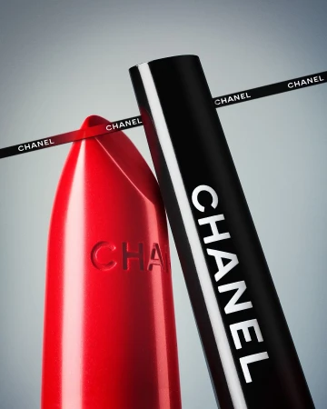 Close-up of a red Chanel lipstick alongside its black cap against a grey gradient background with branded ribbon
