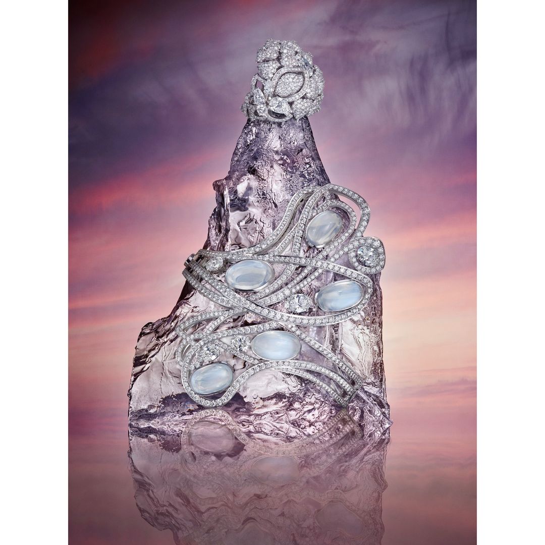 Elevated eye-level shot of diamond jewelry set with moonstones, arranged artistically atop a reflective rock formation against a pink sky.