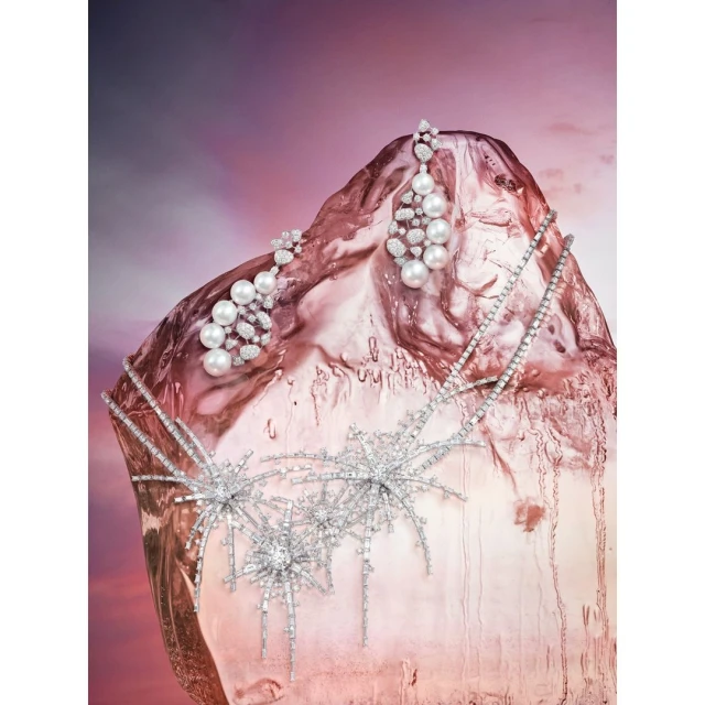 Pearl and diamond earrings and necklace displayed on a textured pink surface against a soft pink background, jewelry product shot.