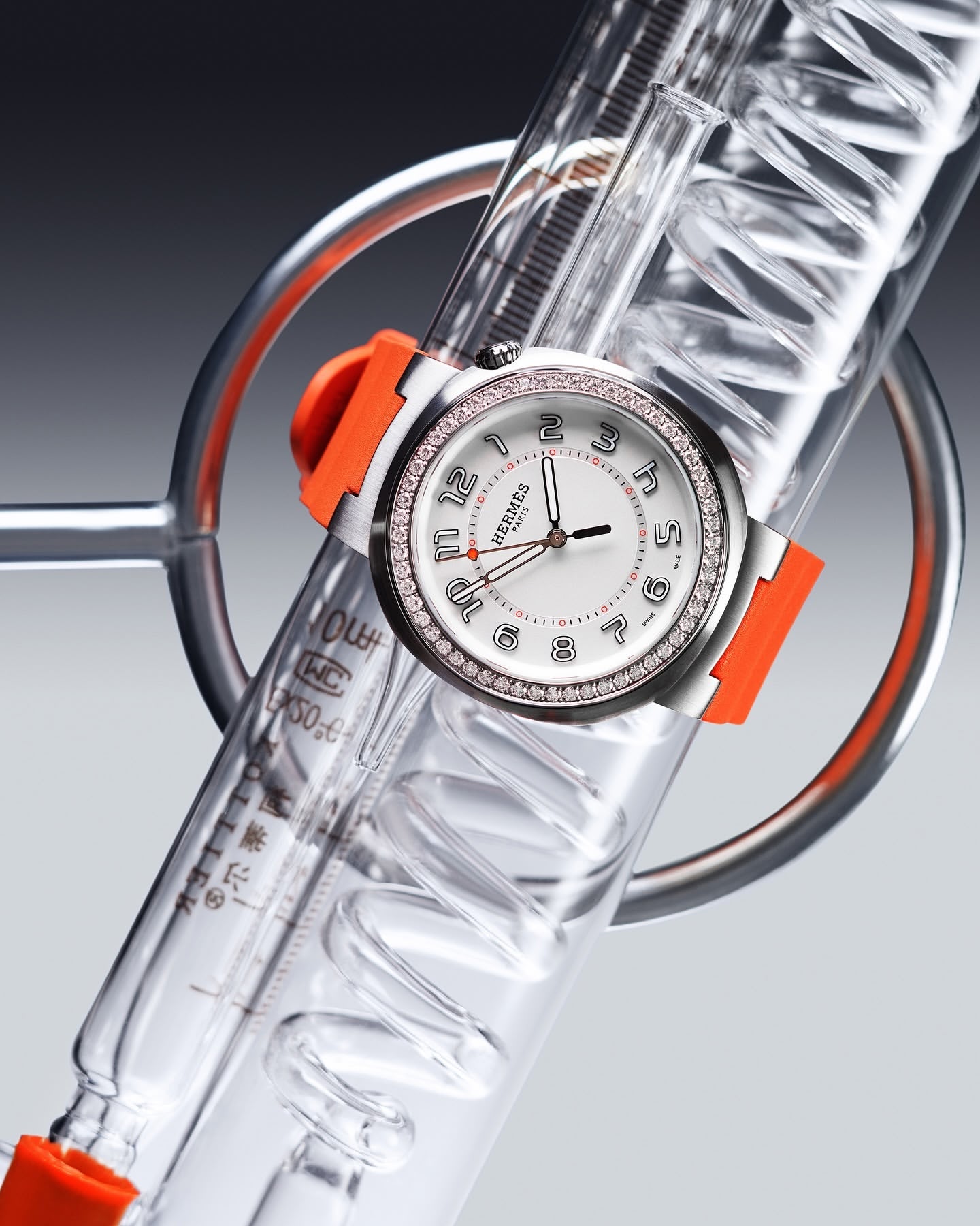 Luxury wristwatch with diamond bezel and orange strap on spiral glass laboratory apparatus against a gradient gray background.