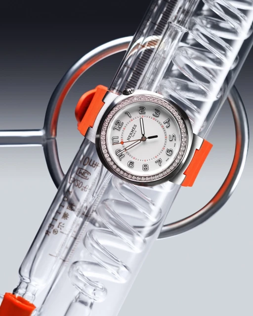 Luxury wristwatch with diamond bezel and orange strap on spiral glass laboratory apparatus against a gradient gray background.