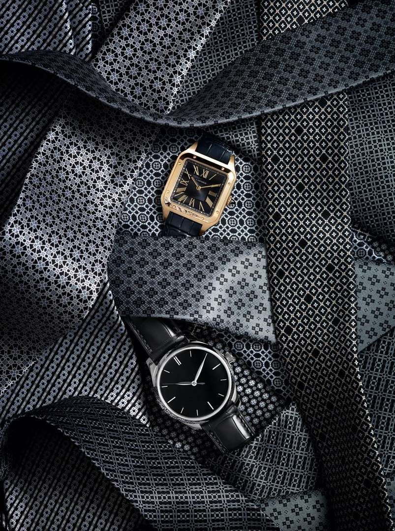 Two luxury wristwatches, one gold-tone and one silver-tone, are showcased amidst a patterned pile of gray and black neckties.