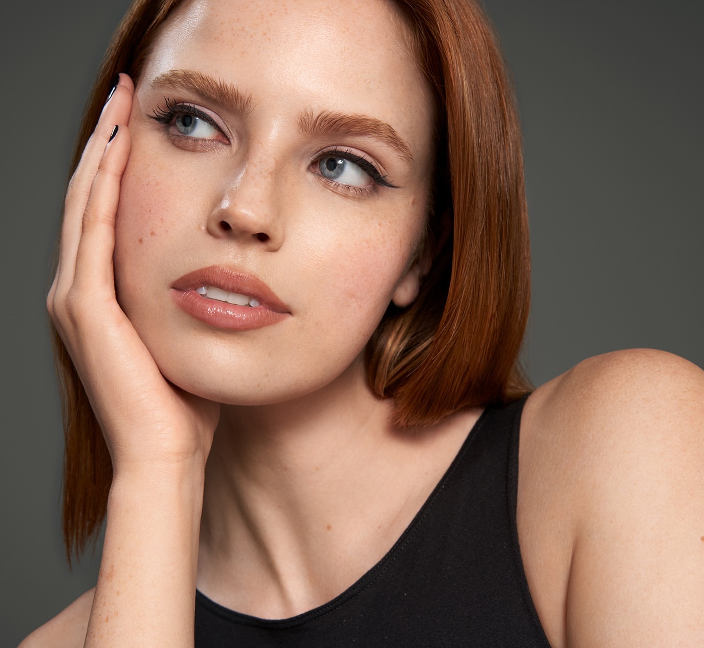 Close-up portrait of a woman with red hair and freckles resting her hand on her cheek against a grey background.