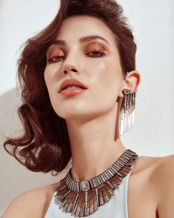 Close-up portrait of a woman wearing an elaborate fringe necklace and statement ear cuff against a plain white wall.