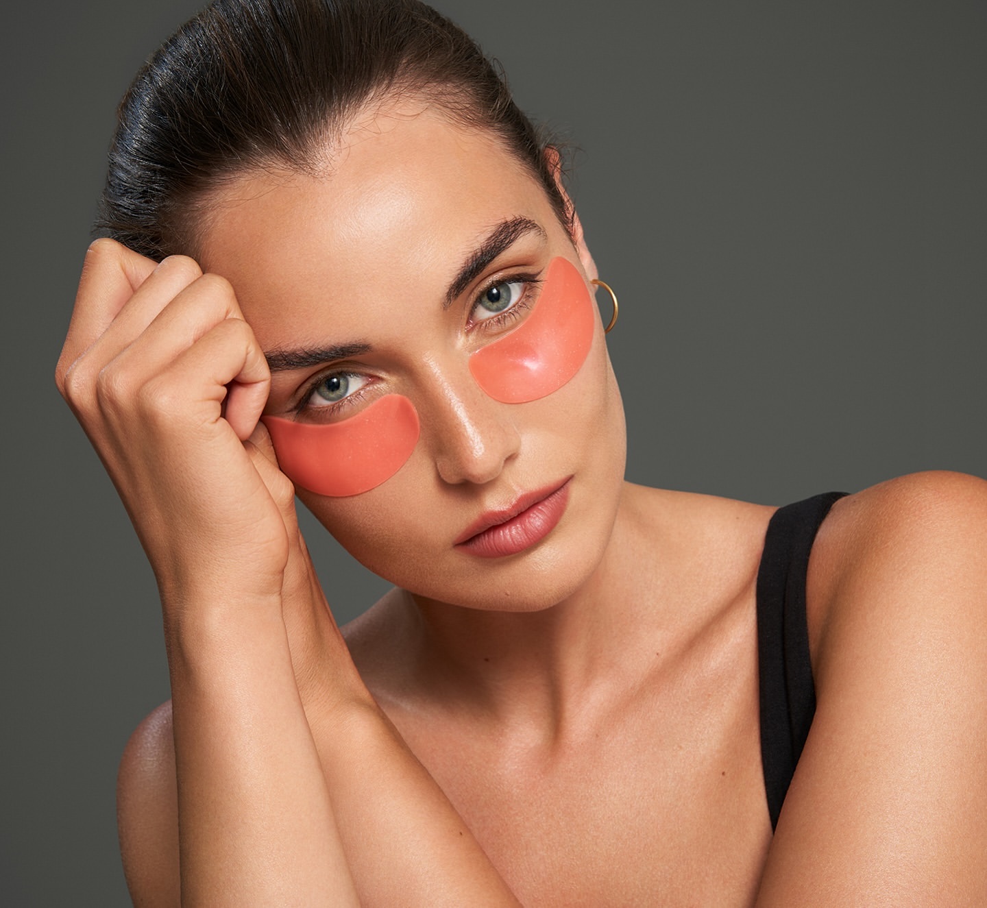 Close-up studio shot of a fair-skinned woman with coral eye patches, wearing a black tank top, resting her head on her hand.