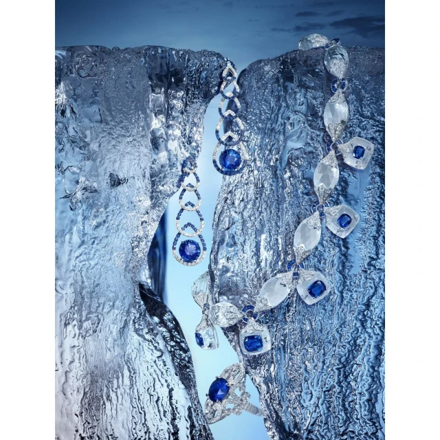 Close-up of sapphire and diamond jewelry, including a necklace, earrings, and ring, displayed on textured ice against a blue background.