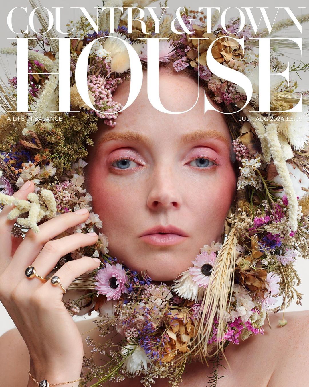 Magazine cover featuring a woman adorned with a flower wreath. The woman gazes directly forward with pink blush and eye shadow.