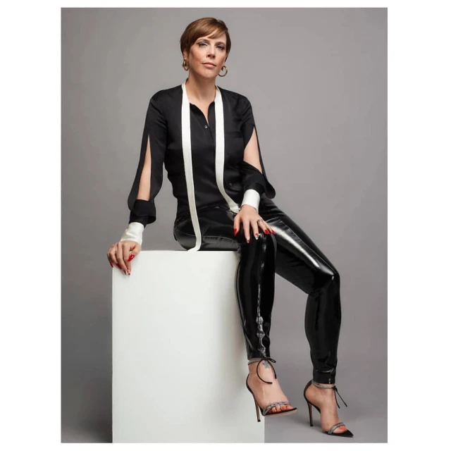 A woman with short hair poses in a black and white blouse and shiny black pants, sitting on a white cube against a grey backdrop.