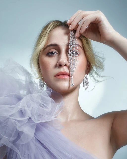 Portrait of a blond woman with fair skin, wearing lilac formal dress, diamond chandelier earrings, and beaded decoration held in hand.