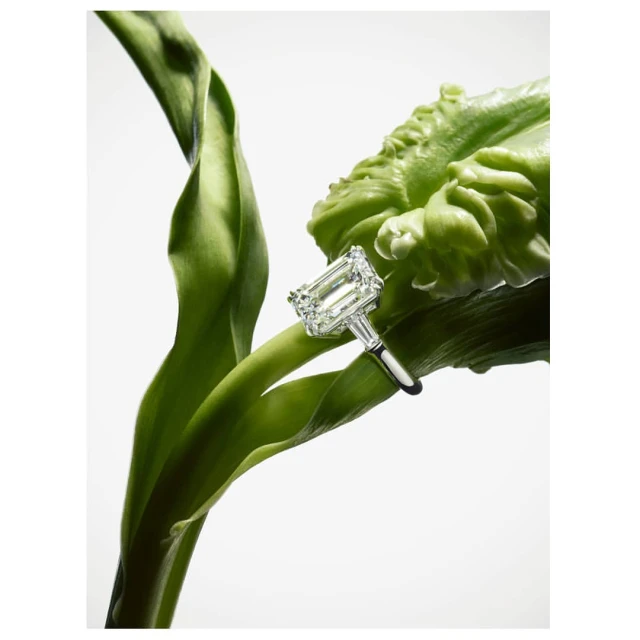 Close-up of a diamond ring on a green flower stem. The ring features an emerald-cut diamond with smaller baguette diamonds.