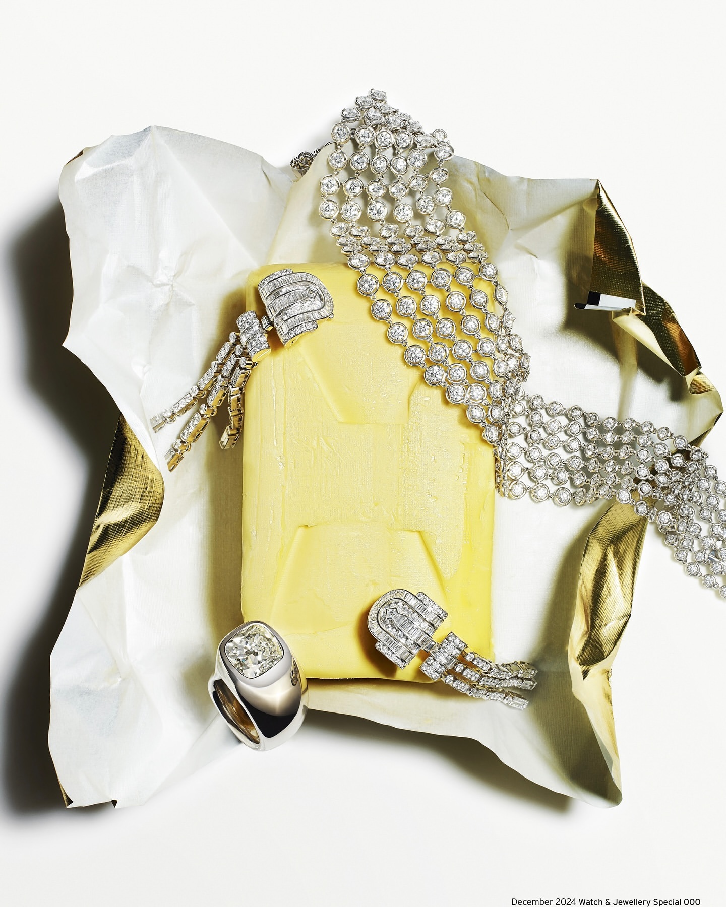 Elevated still life shot of a diamond ring, diamond necklace, and earrings presented on rumpled gold and white paper.