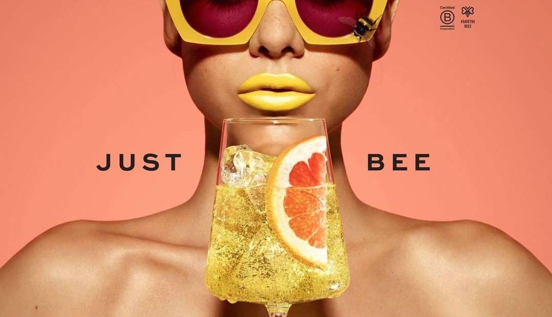 Woman with yellow sunglasses, lips, and a bee on her glasses holds a glass of yellow drink with an orange slice against peach background.