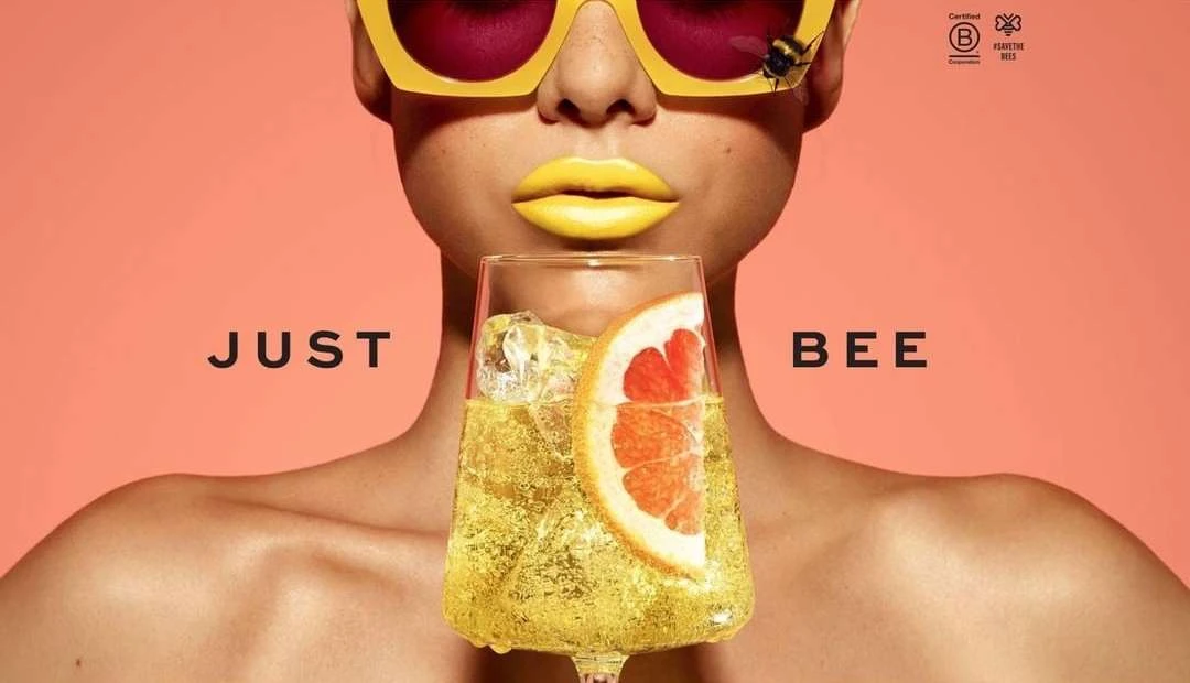 Woman with yellow sunglasses, lips, and a bee on her glasses holds a glass of yellow drink with an orange slice against peach background.