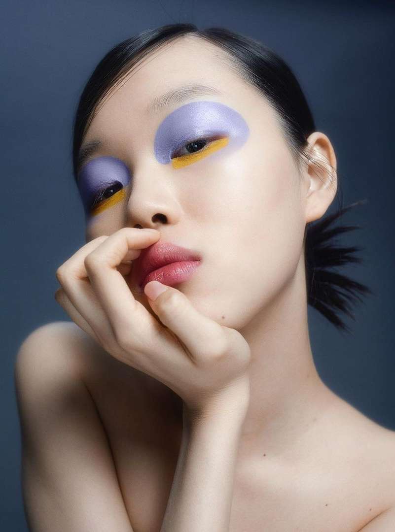 Portrait of an Asian female model with creative eye makeup, featuring lavender, gold, and pink, against a dark gradient background in studio.