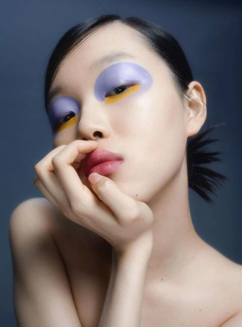 Portrait of an Asian female model with creative eye makeup, featuring lavender, gold, and pink, against a dark gradient background in studio.