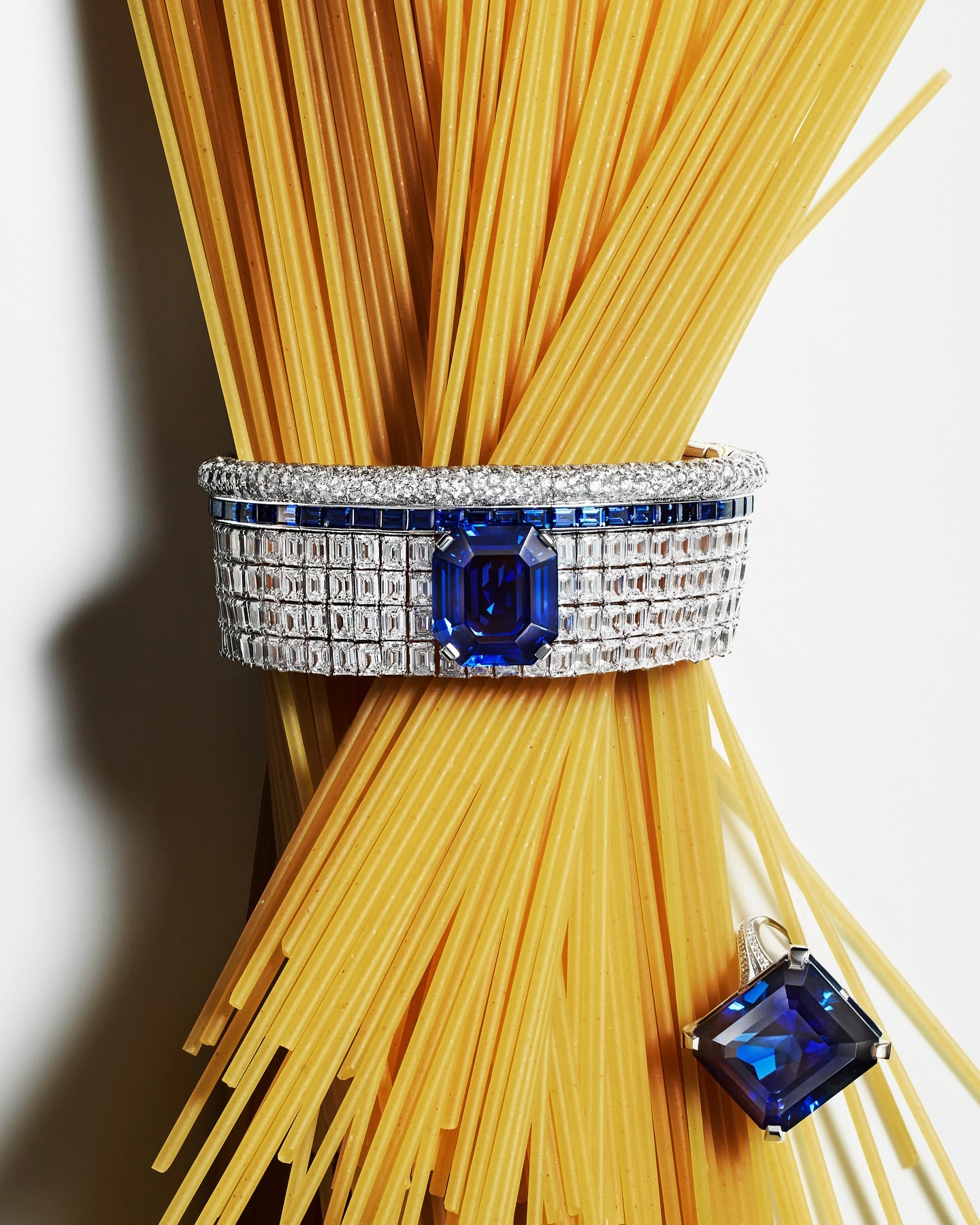 Still life shot of uncooked spaghetti tied with a diamond and sapphire bracelet and matching pendant on a white background.