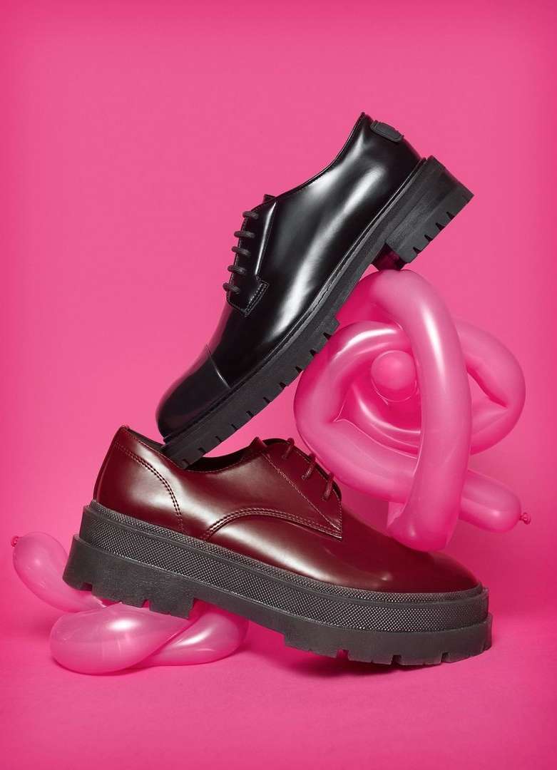 Two platform oxford shoes, one black and one burgundy, are stacked on pink balloon animals, set against a vibrant pink backdrop.