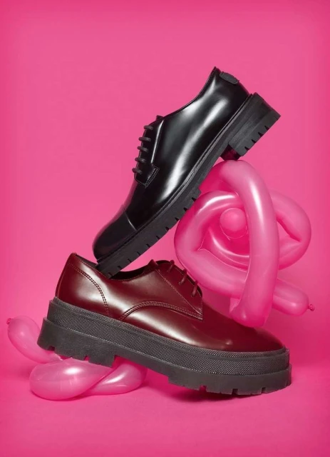 Two platform oxford shoes, one black and one burgundy, are stacked on pink balloon animals, set against a vibrant pink backdrop.