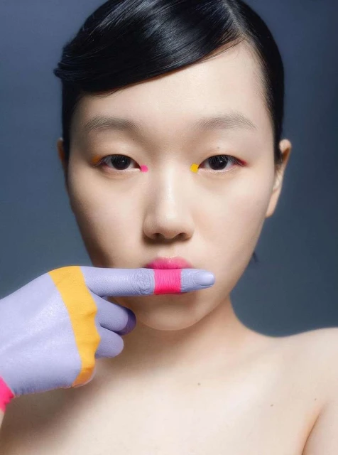 Asian woman's close-up portrait with lilac painted hand covering mouth, colorful face makeup, dark hair, gray studio background.