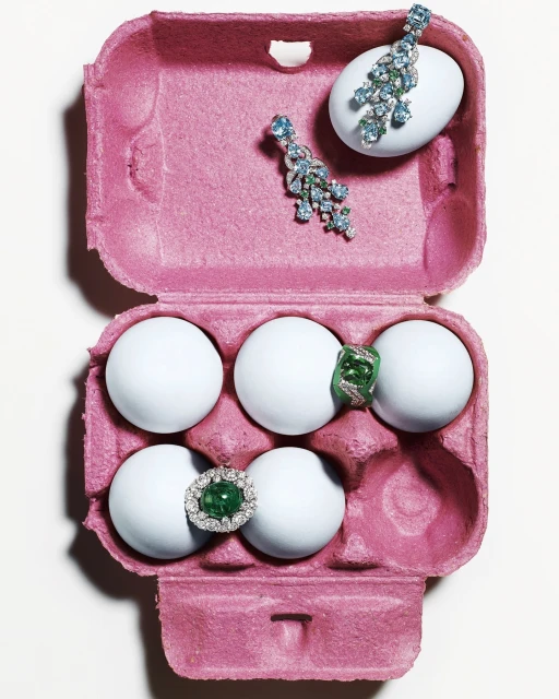 Overhead studio shot: gemstones and jewelry on white eggs inside a pink egg carton, creating an eye-catching, colorful composition.