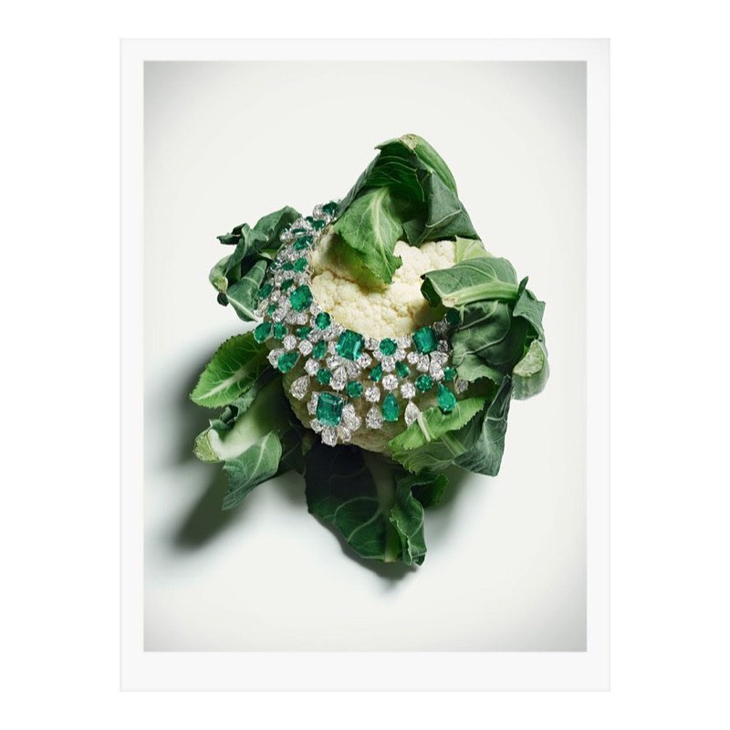 High-angle shot of a diamond and emerald necklace draped over a white cauliflower, set against a bright white background for a modern jewelry advertisement.