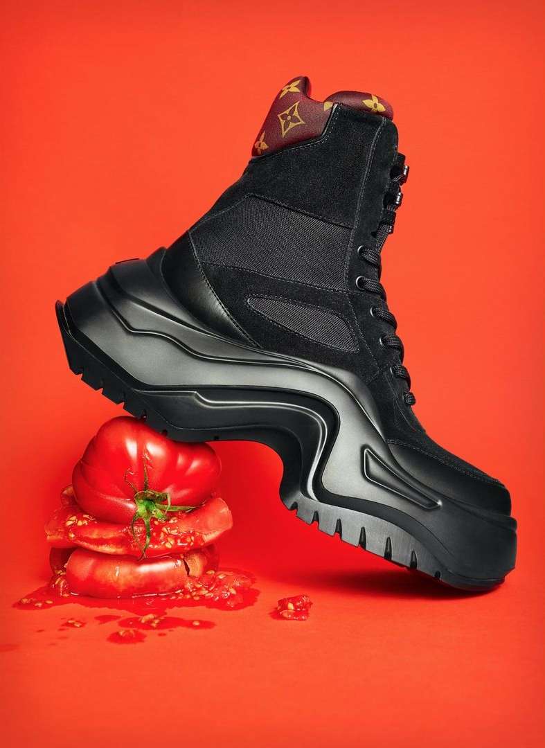 Black combat boot with a thick lug sole is placed on a crushed pile of tomatoes against an orange studio backdrop.