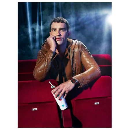 Adult man in a brown leather jacket sits in a red theatre seat, illuminated by a spotlight in a studio shot.