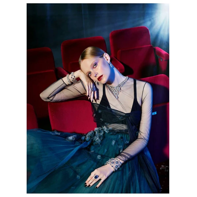 A woman models jewelry in a dark theater with red plush seats, wearing a black and teal dress, necklace, earrings, rings, and bracelets.