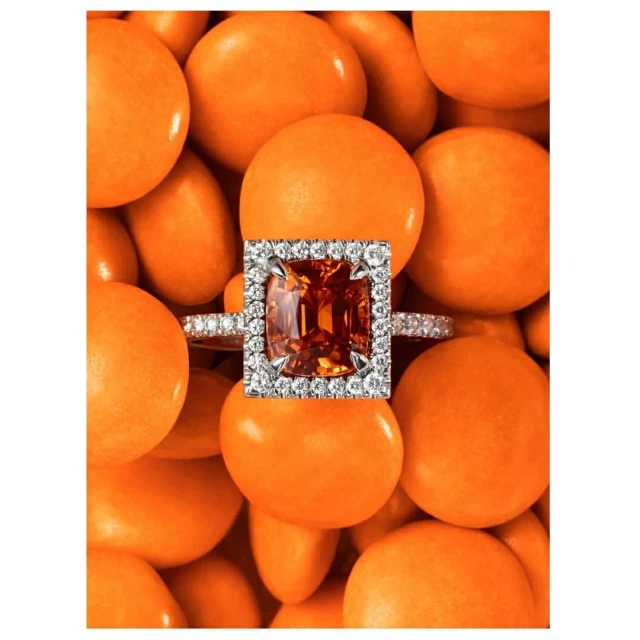 Close-up of a silver engagement ring featuring a vibrant orange gemstone surrounded by diamonds, set against a background of orange candies.
