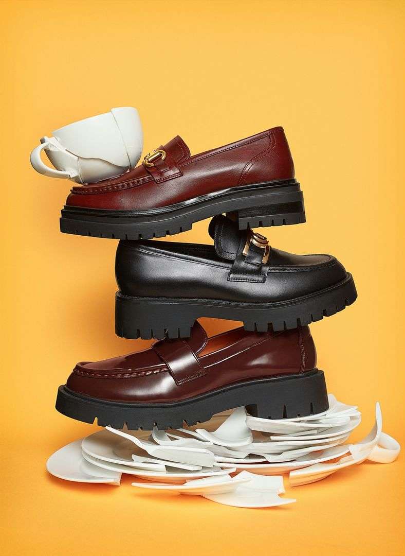 Stacked leather loafers in red and black, balanced with a white teacup on top and shattered white plates at the base, set against a yellow backdrop.