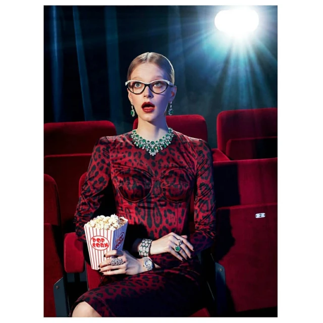 Woman in animal-print dress with jewelry seated in red theatre, eating popcorn under bright spotlight, with dark teal and red background.