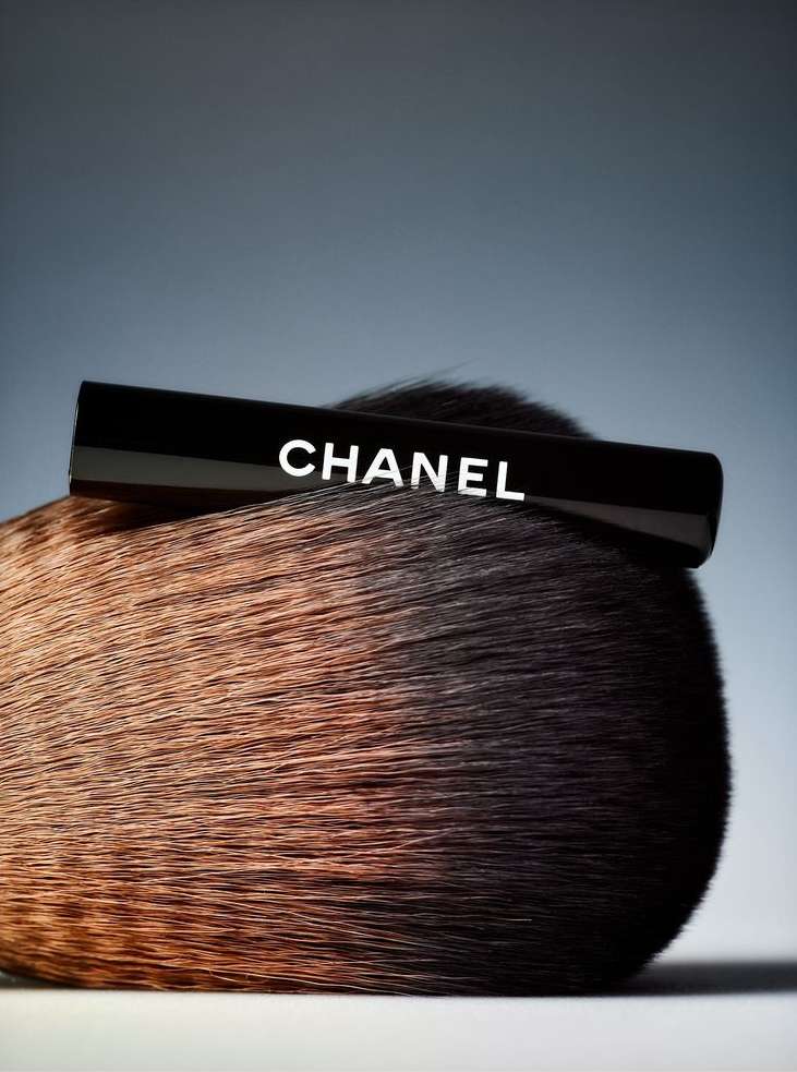 Chanel brand make-up brush with black handle and soft brown bristles on gradient grey background in beauty product photography.