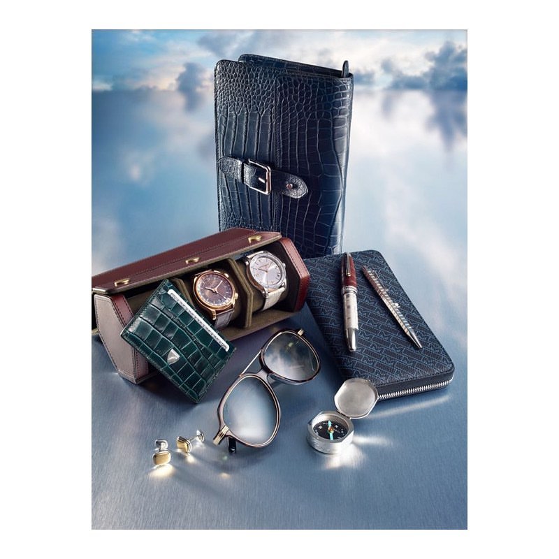 Elevated shot showcasing luxury accessories: watches in a case, crocodile wallet, pens, eyeglasses, and a compass against a cloudy sky.