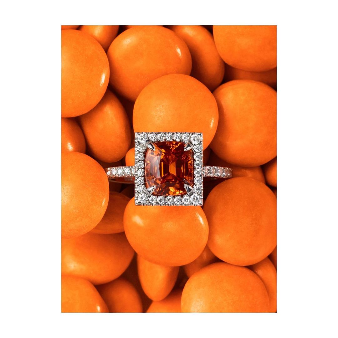 Close-up of a silver ring with an orange gemstone surrounded by diamonds, set against a background of orange candy spheres.
