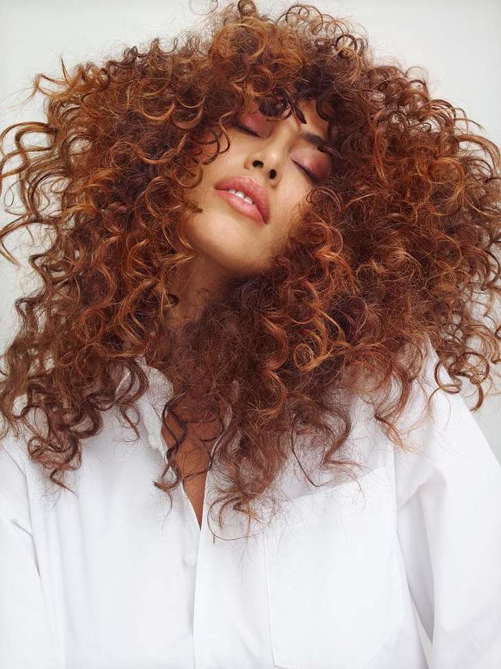 Close-up portrait of a woman with voluminous curly auburn hair and eyes closed wearing a white button-down shirt.