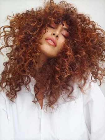 Close-up portrait of a woman with voluminous curly auburn hair and eyes closed wearing a white button-down shirt.
