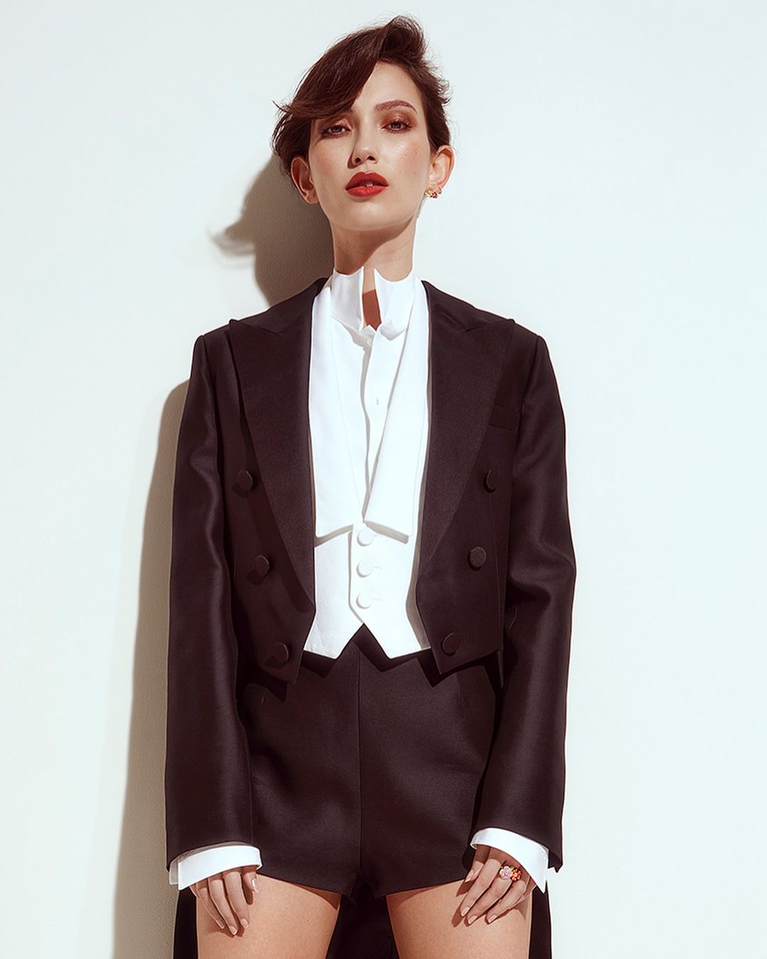 Elegant woman poses in a black tuxedo jacket and shorts against a white background, showcasing a modern take on classic formalwear.