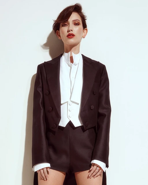 Elegant woman poses in a black tuxedo jacket and shorts against a white background, showcasing a modern take on classic formalwear.