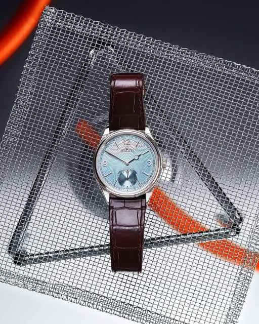 Wristwatch on a textured metal surface, close-up. Features include a light blue dial, brown leather strap, and silver casing.
