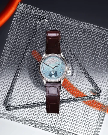 Wristwatch on a textured metal surface, close-up. Features include a light blue dial, brown leather strap, and silver casing.