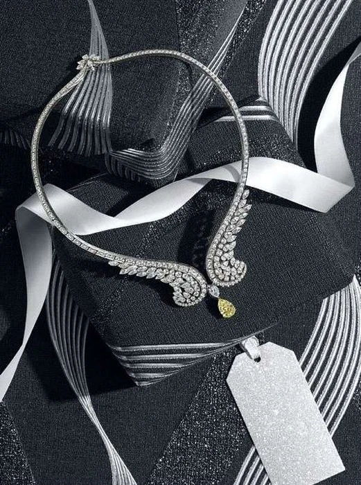 Diamond necklace with wing motifs and a yellow drop stone resting on textured dark packages tied with white satin ribbons.