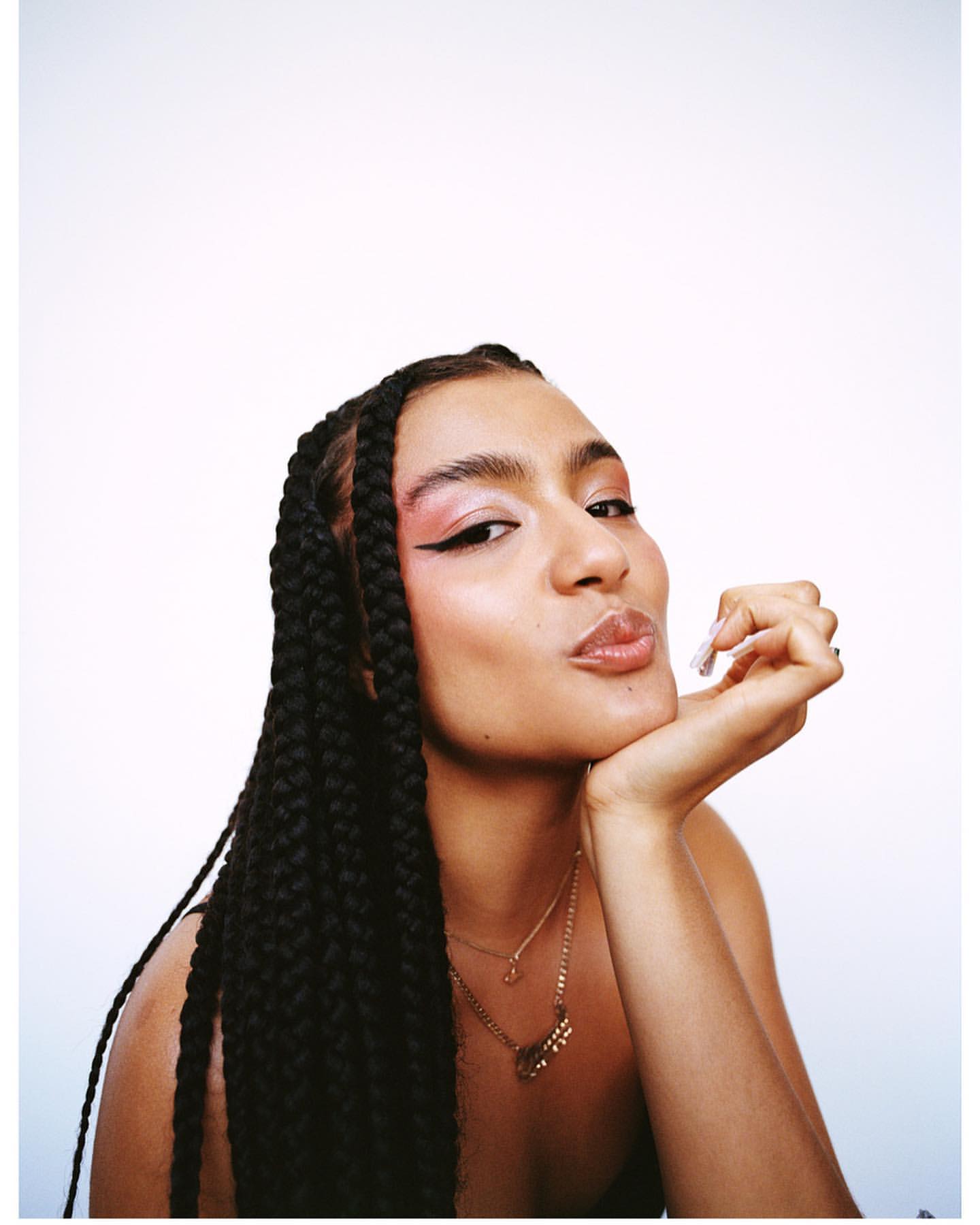 Portrait of a woman with braided hair, wearing pink eyeshadow and a gold necklace, pouting in front of a plain white background.