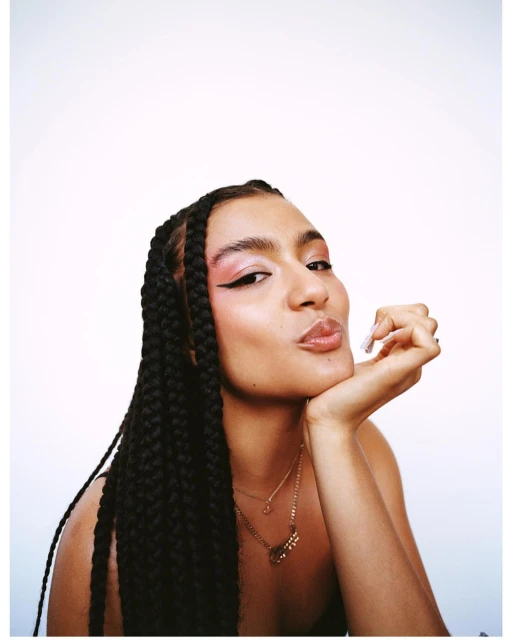 Portrait of a woman with braided hair, wearing pink eyeshadow and a gold necklace, pouting in front of a plain white background.